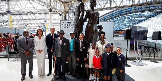 El príncipe William, Catherine, duquesa de Cambridge, la baronesa Floella Benjamin, a la derecha, los pasajeros de Windrush, Alford Gardner, al centro, John Richards, cuarto desde la izquierda, y los niños en el Monumento Nacional de Windrush creado por el artista jamaicano Basil Watson, a la izquierda, en la estación de Waterloo en Londres El 22 de junio de 2022.