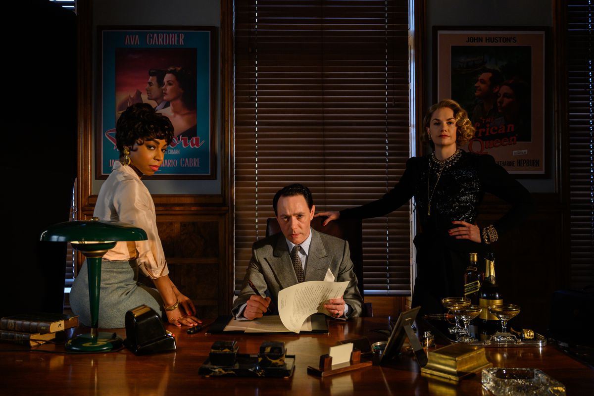 John Wolfe (Rice Shersmith), Petola Spencer (Rita Wilson) y Anne Savile (Peppa Bennett Warner) miran desde detrás del escritorio grande y brillante de Wolfe.