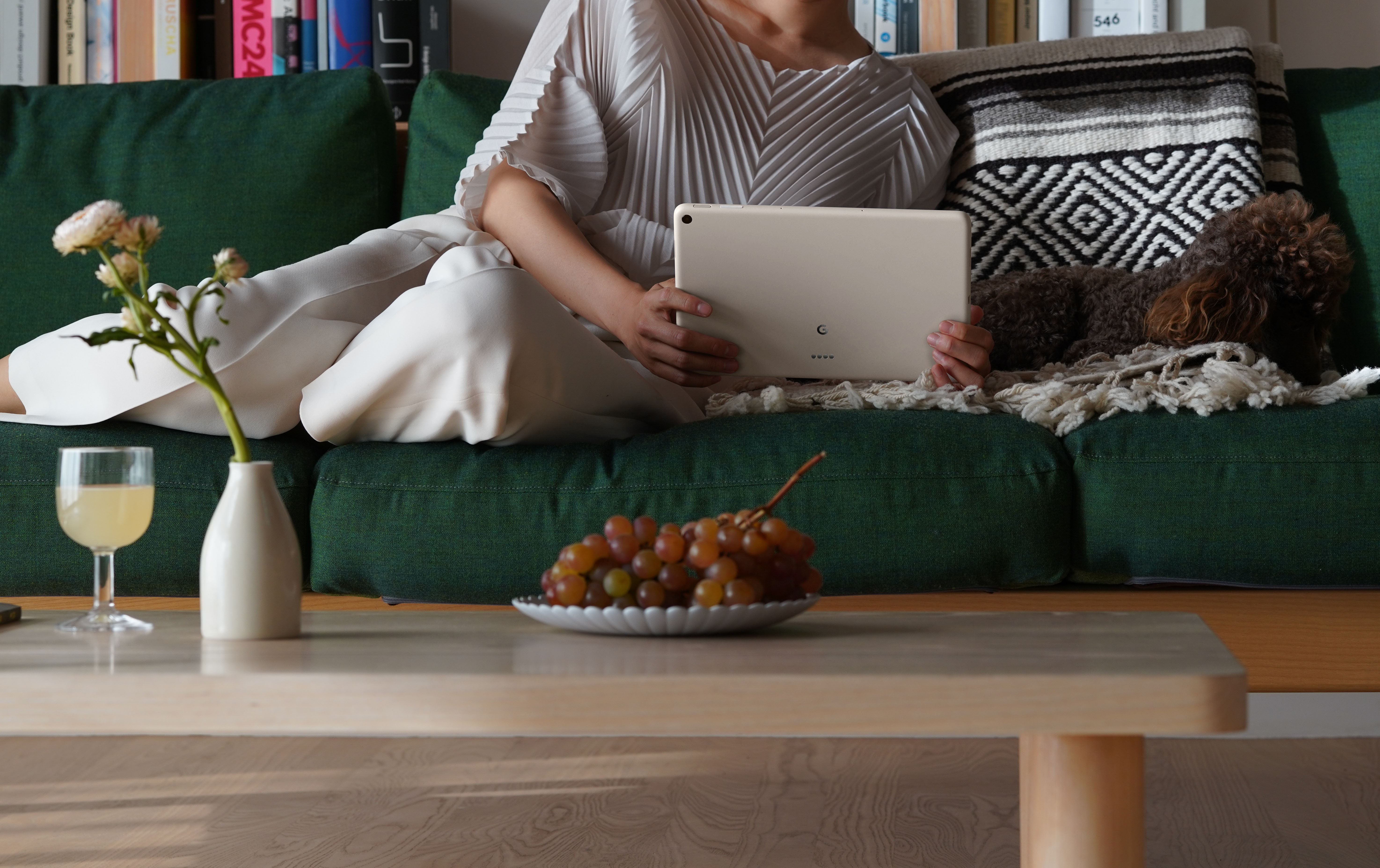 A person holding a white Pixel Tablet while laying on a green couch with a plate of grapes on a table in front of them. The back of the Tablet is facing out and there is a camera on the top left of the device's rear.